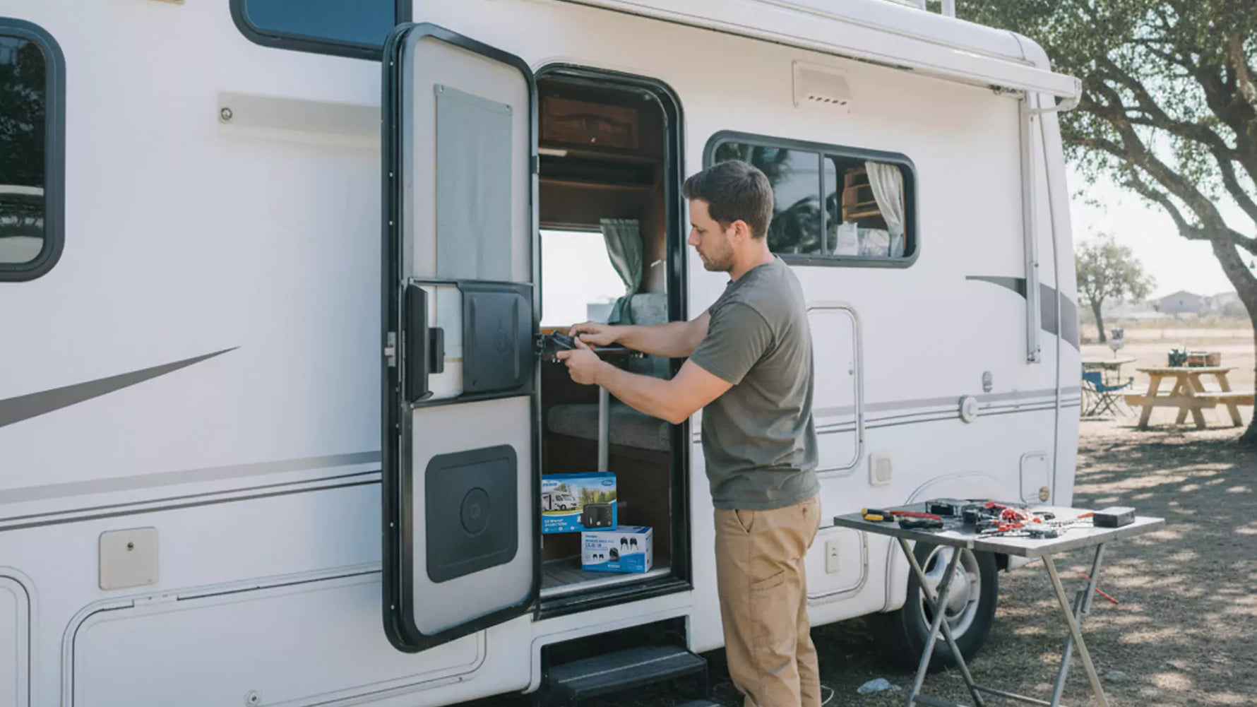 A man stands outside an RV fixing the door lock, with tools and boxes on a nearby table in a campground setting.