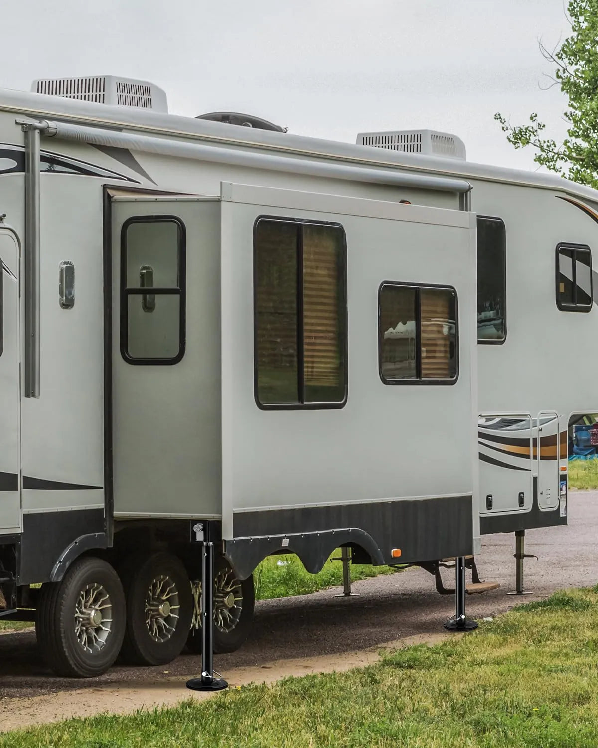 A light gray and black travel trailer parked outdoors on grass and dirt, with the side door open and support legs extended.